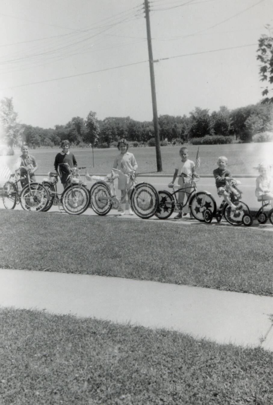 Black and white snapshot of a group of children with decorated bikes. They are outside on a street.